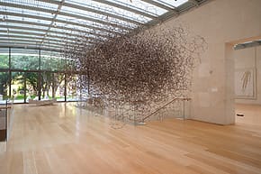 Large intricate metallic sculpture suspended in a spacious, sunlit gallery with wooden floors and glass walls.