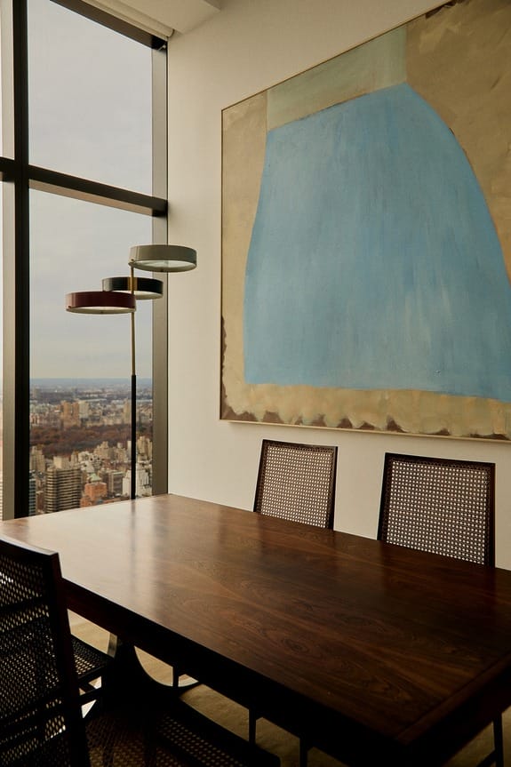 Modern dining room with a wooden table, chairs, abstract art on the wall, and a view of the city through large windows.
