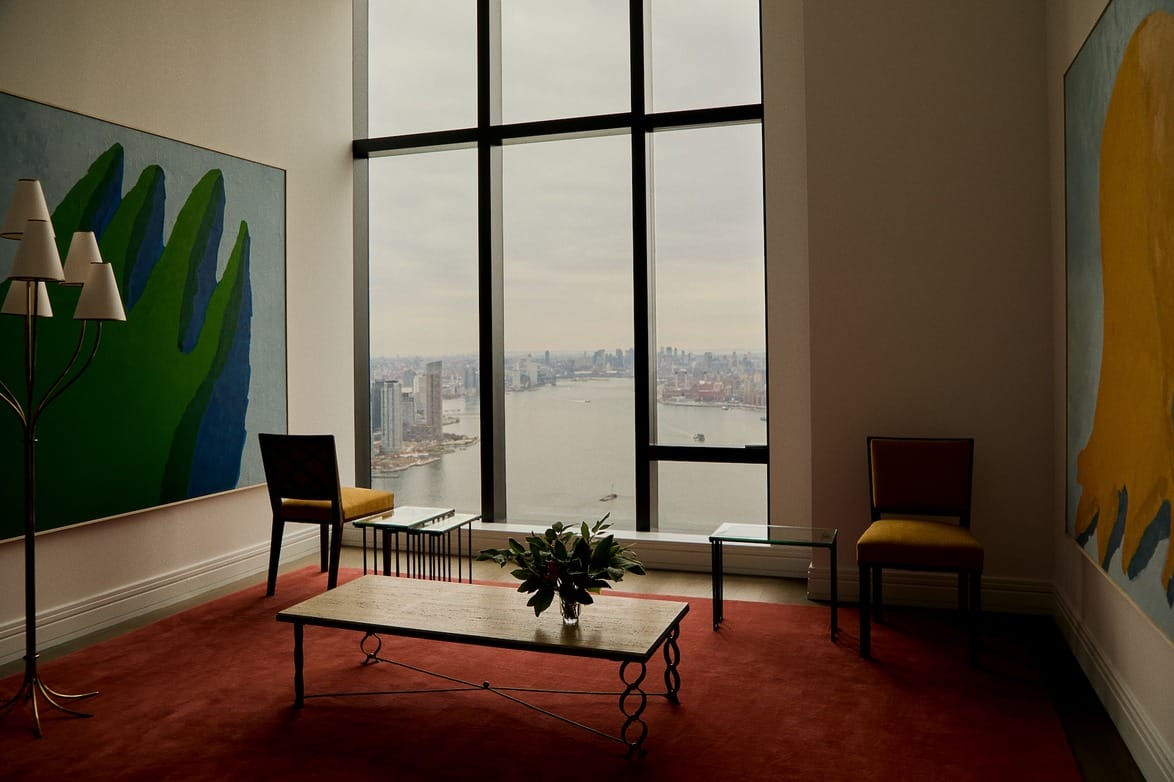 Room with large window, cityscape view, artwork on wall, red rug, chairs, and a table with a plant centerpiece.