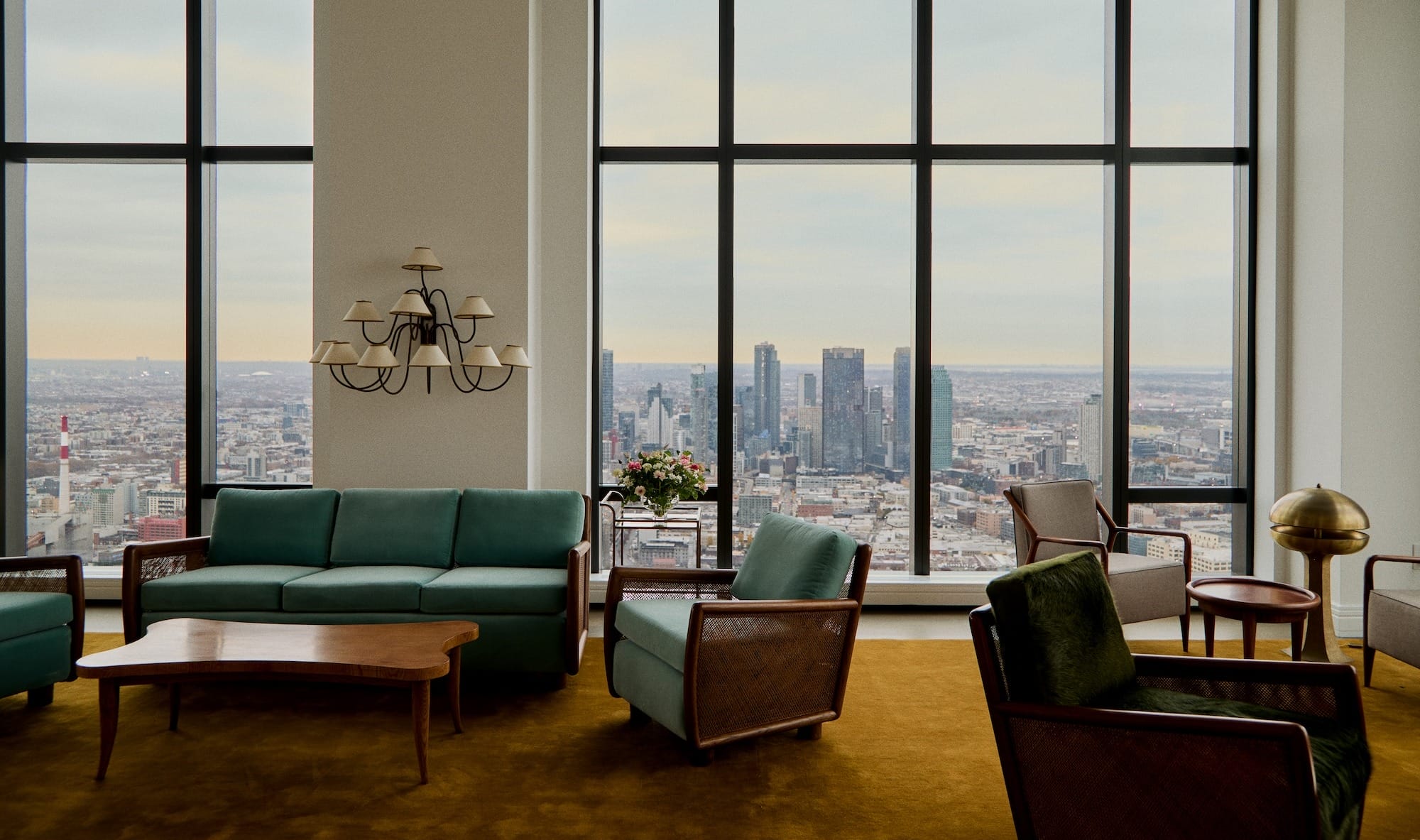 Luxurious hotel lounge with green sofas, large windows offering a city skyline view, and a chandelier hanging above.