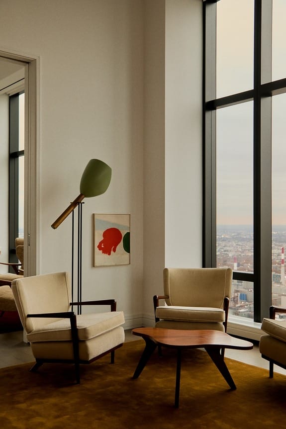 Modern living room with large windows, beige armchairs, abstract art, and a floor lamp, overlooking a cityscape.