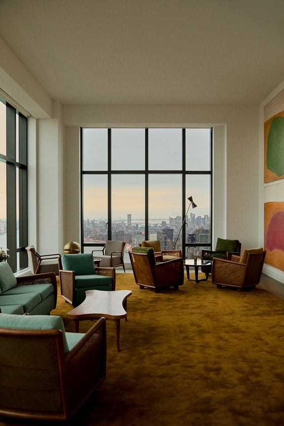 Spacious living room with retro furniture, large windows, and a view of the city skyline at sunset.
