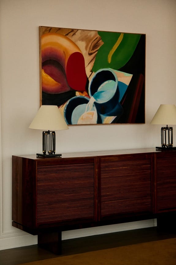 Modern wooden sideboard with two table lamps, set against a colorful abstract painting on a white wall.