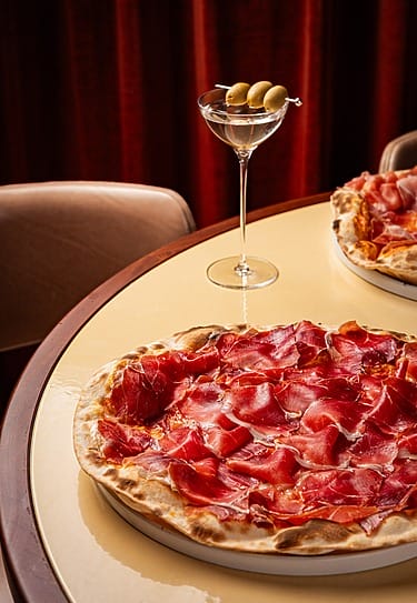 Pizza topped with thinly sliced prosciutto on a table next to a martini glass with olives in a cozy dining setting.