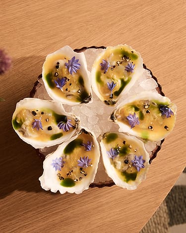 Six oysters on a bed of ice, garnished with vibrant purple flowers and a green sauce, served on a wooden table.