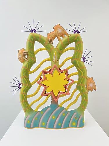 Colorful abstract sculpture with a central star shape, green wavy lines, orange hands, and purple starbursts on top.