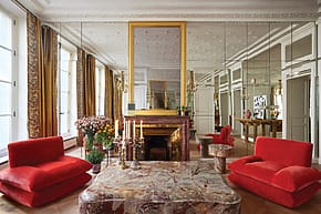 Luxurious living room with mirrored walls, ornate fireplace, vibrant red sofas, and a large marble coffee table.