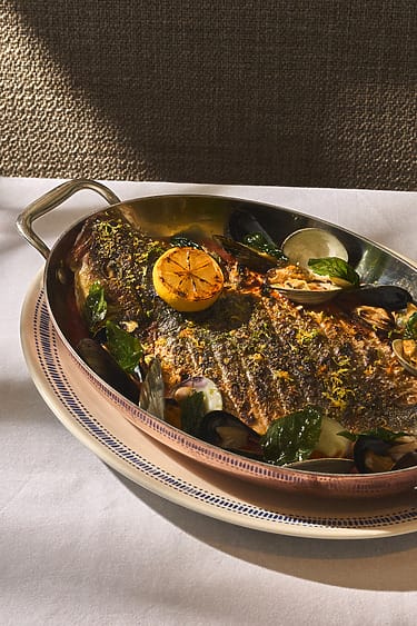 Grilled fish garnished with lemon, herbs, and mussels on a metal platter placed on a white tablecloth.