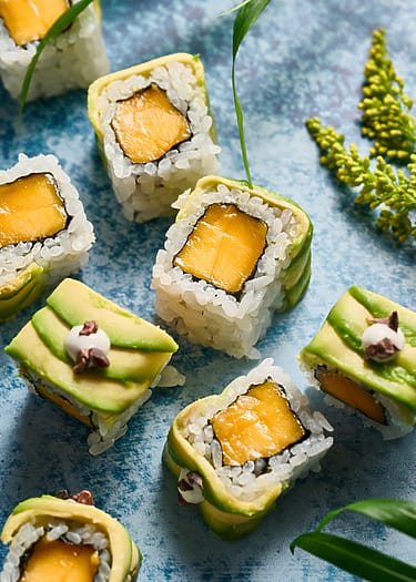 Sushi rolls with mango and avocado on a textured blue surface with green leaves and a sprig of yellow flowers.