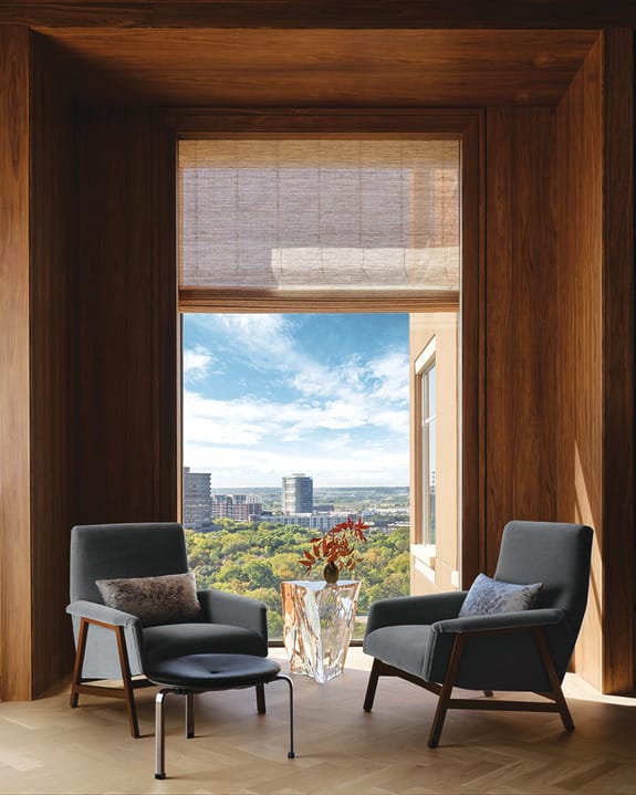 Two armchairs facing each other in a wooden room with a cityscape view and blue sky visible through a large window.