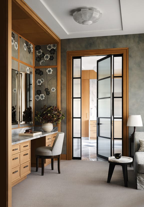 Elegant home office with wooden desk, chair, large mirror, floral wallpaper, and glass-paneled doors opening to another room.