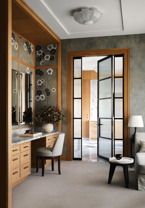 Elegant home office with wooden desk, chair, large mirror, floral wallpaper, and glass-paneled doors opening to another room.