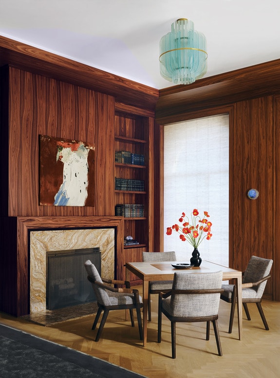 Elegant dining room with wooden walls, marble fireplace, chandelier, table set for four, and painting on wall.
