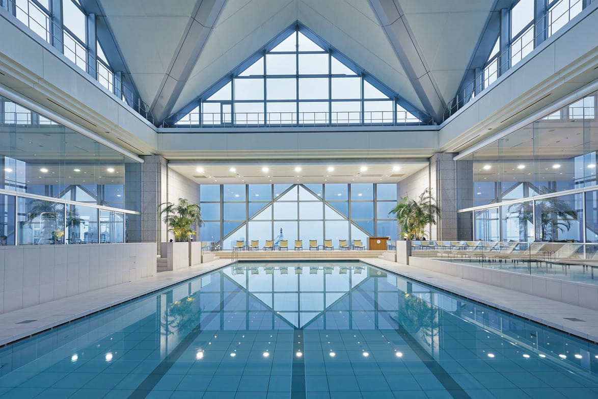 Indoor swimming pool with symmetrical architecture and large windows, surrounded by lounge chairs and potted plants.