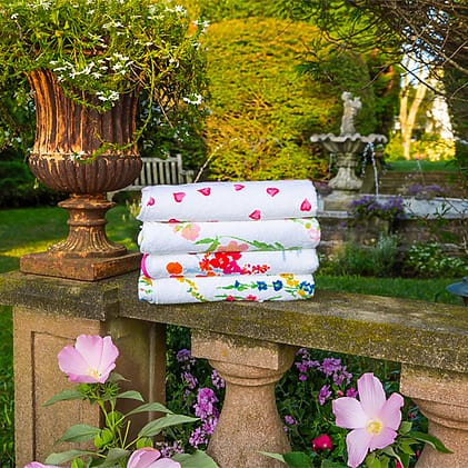Colorful towels stacked on a stone railing in a garden with flowers, a fountain, and a decorative planter nearby.