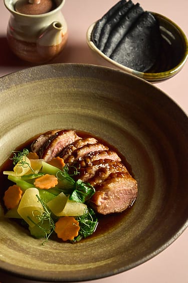 Sliced duck breast with sauce, garnished with vegetables on a ceramic plate, served with black tortillas and a clay pot.