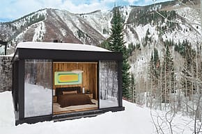 Modern cabin with glass walls in snowy mountain landscape, featuring a cozy bedroom and artistic decor.