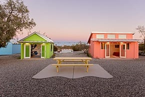 Colorful tiny houses with a yellow picnic table in between, set against a scenic sunset sky and gravel foreground.
