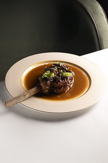 Braised beef rib served on a plate with rich sauce and garnished with fresh herbs, placed on a white table.