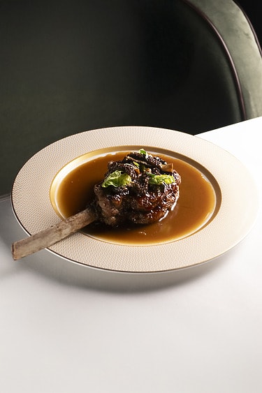 Braised beef rib served on a plate with rich sauce and garnished with fresh herbs, placed on a white table.