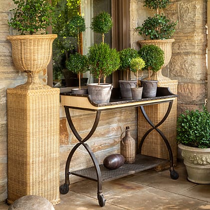 Outdoor patio with potted plants on a rustic metal cart, flanked by two tall wicker planters, against a stone wall.