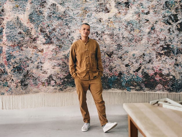 Person in brown outfit stands in front of abstract art wall, leaning casually, with hands in pockets, in a bright room.
