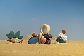 Abstract arrangement of various objects on a sandy surface with pyramids and clear blue sky in the background.