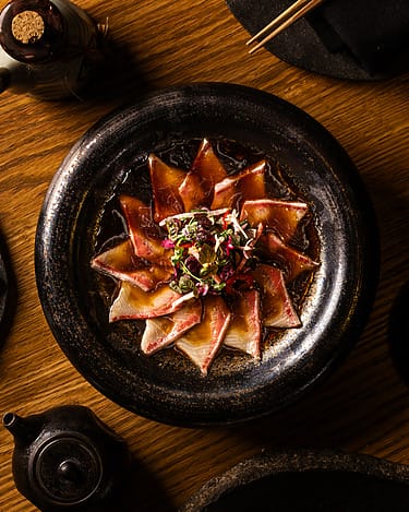 Dish with artfully arranged slices of fish topped with microgreens on a dark plate, set on a wooden table with teapot nearby.