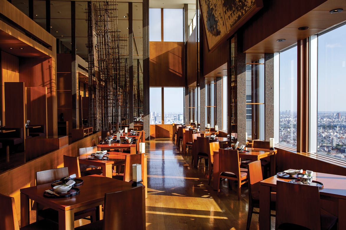 Modern restaurant interior with large windows, wooden tables, and panoramic city view.