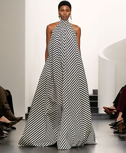 Model on runway wearing a flowing black and white striped dress with high neckline, in a fashionable setting.