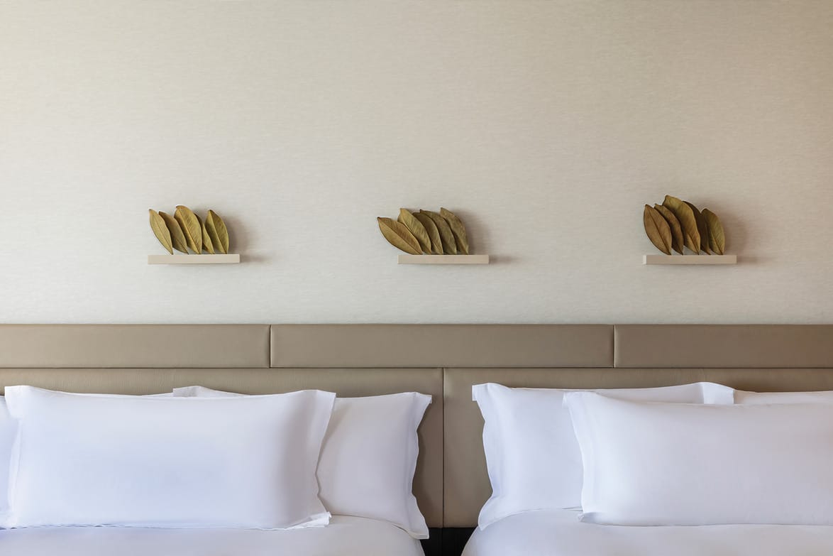 Elegant hotel room with two neatly made beds, white linens, and three decorative leaf sculptures on a beige wall above the headboards.