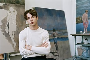 Young man sitting in an art studio with folded arms, surrounded by coastal-themed paintings in various stages of completion.