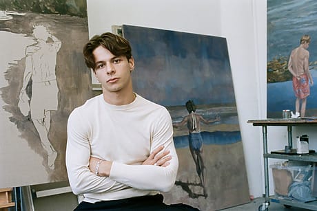 Young man sitting in an art studio with folded arms, surrounded by coastal-themed paintings in various stages of completion.