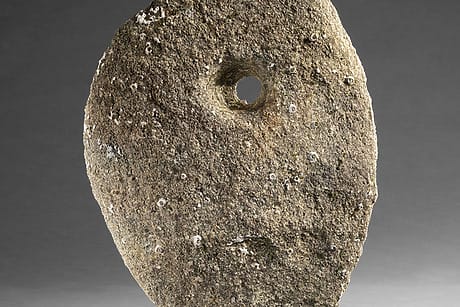 Oval stone with circular hole in the center, displayed on a dark base against a gray background.