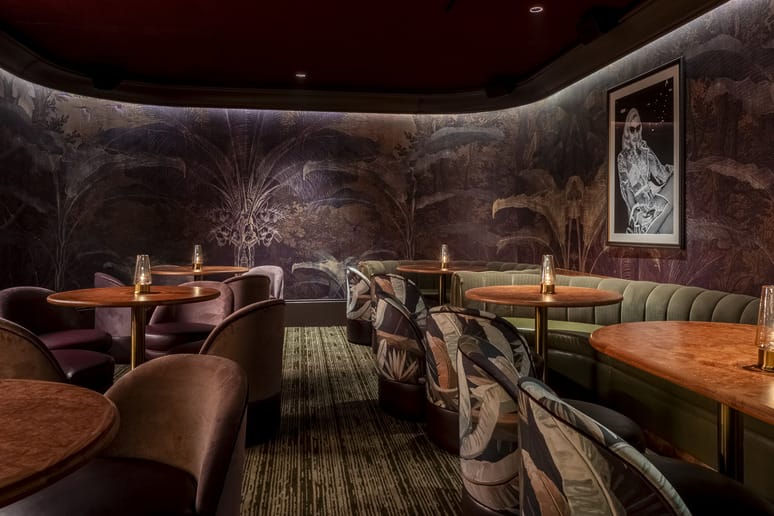 Cozy, dimly lit lounge with elegant seating, tropical-themed wallpaper, and small lanterns on round wooden tables.