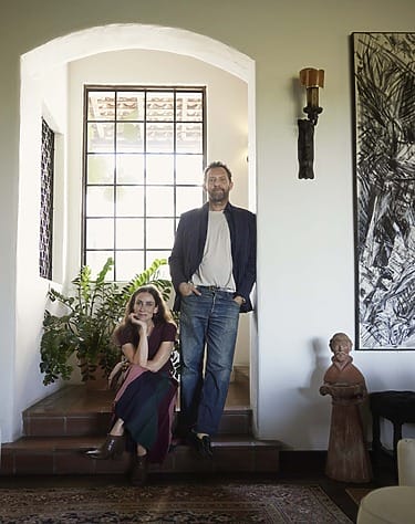Man and woman posing together in a sunlit room with large window, plants, and artwork in background.