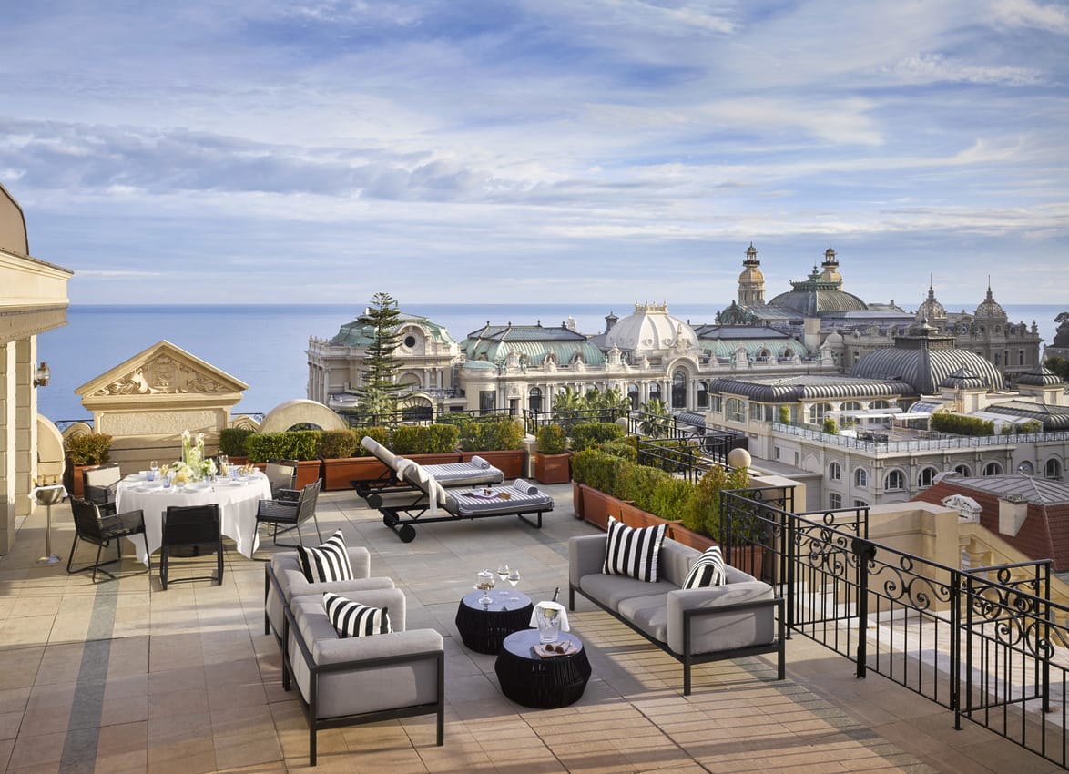 Luxurious rooftop terrace with elegant furniture, overlooking cityscape and sea under a clear blue sky.