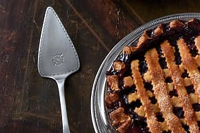 Pie server next to a partially eaten berry pie with a lattice crust on a dark wooden table.