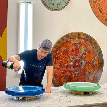 Artist using a blowtorch to create colorful circular patterns on two large ceramic plates in a studio setting.