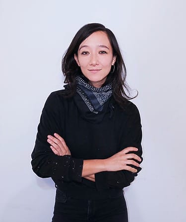 Person with dark hair wearing a black sweater and scarf, standing against a light background with arms crossed.