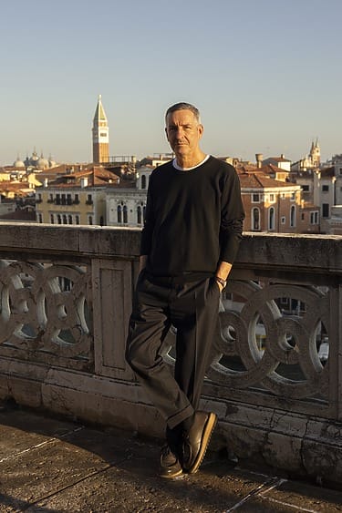 Man standing on balcony overlooking cityscape with historic buildings and bell tower in background during sunset.