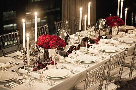 Elegant dinner table setup with white linens, red floral centerpieces, candles, and silverware in a dimly lit room.