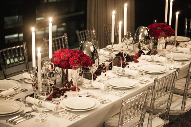 Elegant dinner table setup with white linens, red floral centerpieces, candles, and silverware in a dimly lit room.