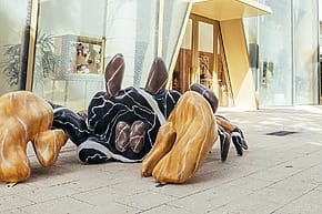 Abstract sculpture with black, white, and brown forms on a city sidewalk near a modern building entrance.