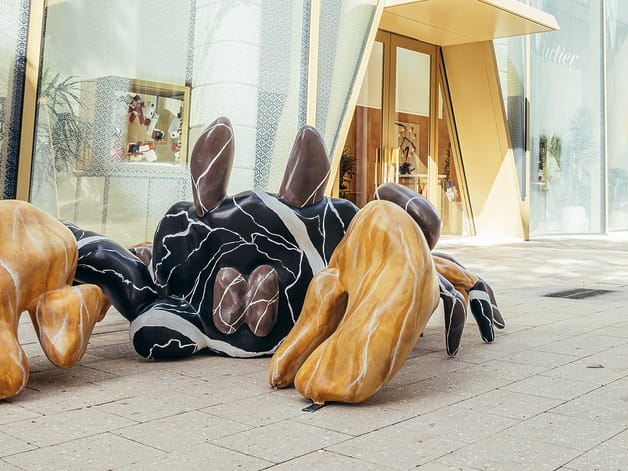 Abstract sculpture with black, white, and brown forms on a city sidewalk near a modern building entrance.