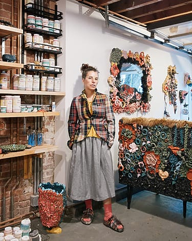 Artist in a studio surrounded by colorful handmade sculptures and shelves filled with art supplies.