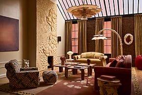Eclectic living room with patterned furniture, carved wall art, large chandelier, and tall windows with brown curtains.