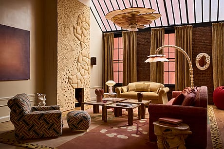 Eclectic living room with patterned furniture, carved wall art, large chandelier, and tall windows with brown curtains.