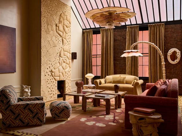 Eclectic living room with patterned furniture, carved wall art, large chandelier, and tall windows with brown curtains.