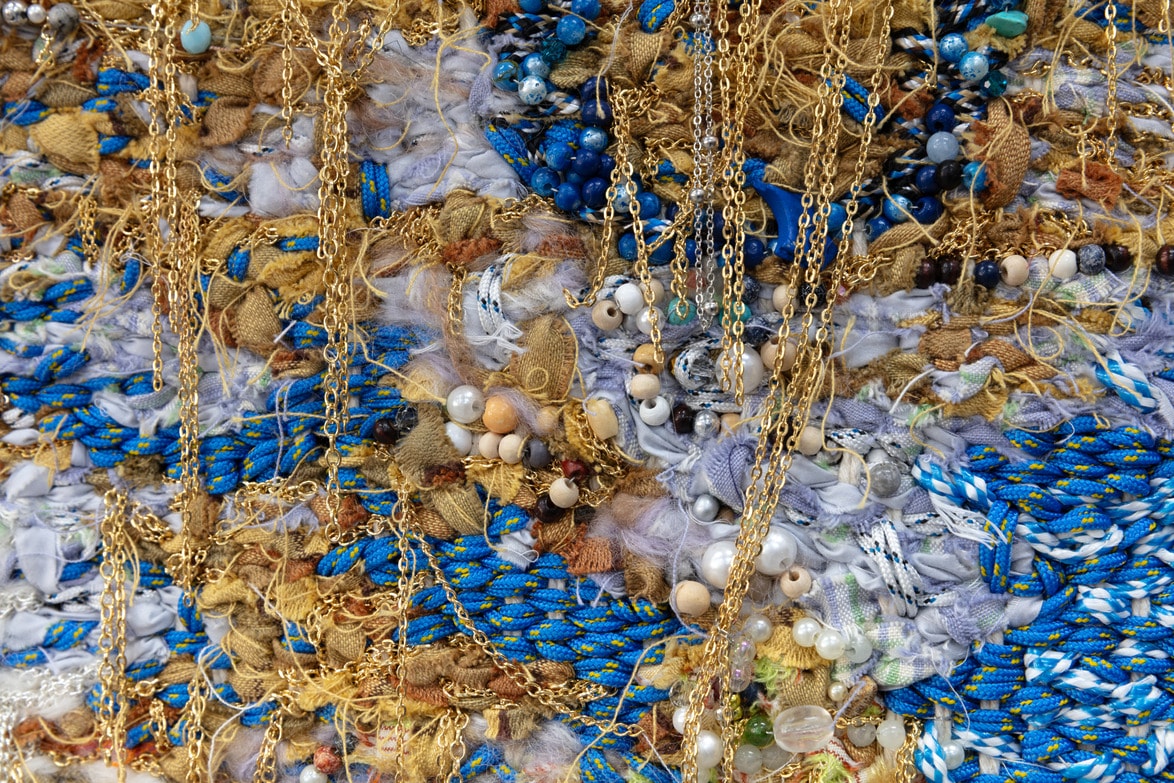 Colorful abstract textile art with blue, gold, and beige hues, featuring chains, beads, and textured fabric layers.